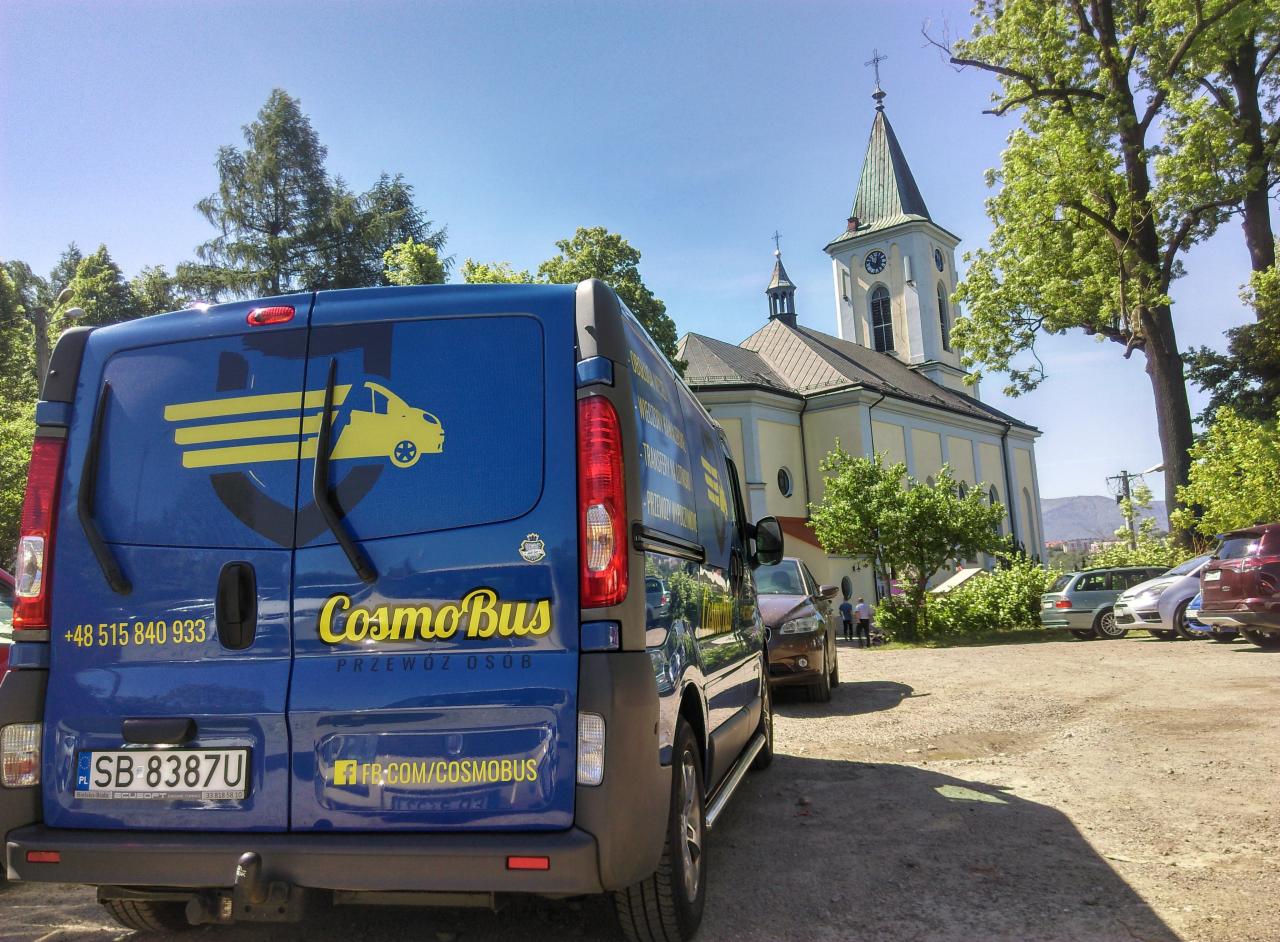 Niebieski bus z logo firmy CosmoBus zaparkowany przed jasnym kościołem z wieżą zegarową, w tle inne samochody i zieleń.