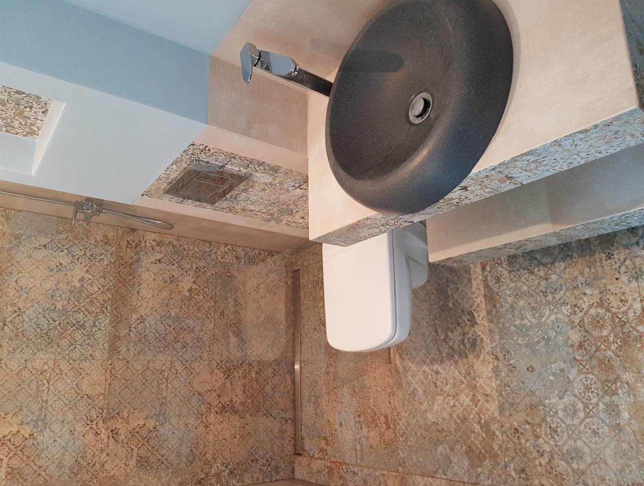 Nowoczesna łazienka z szarą, okrągłą umywalką nablatową i podwieszaną toaletą, wykończona płytkami w odcieniach beżu i brązu z delikatnym wzorem.