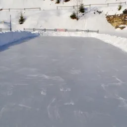 Zimowa sceneria z przygotowanym lodowiskiem otoczonym śnieżnymi zaspami i drewnianą budką w tle.