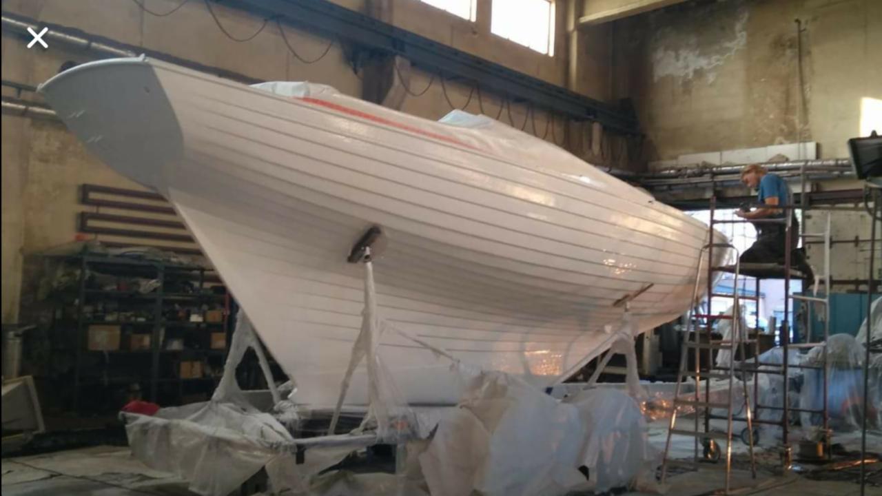 Biały kadłub łodzi na stojakach w hali warsztatowej, pracownik na drabinie wykonuje prace wykończeniowe.