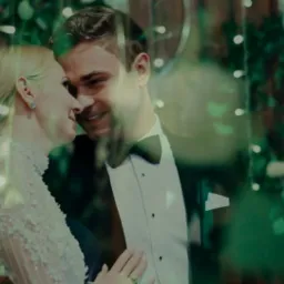 Uśmiechnięta para młoda, panna młoda w koronkowej sukni z kwiatowymi aplikacjami i delikatnym upięciu, pan młody w smokingu, w tle dekoracja z zielonych gałązek i światełek, ujęcie przez szybę.