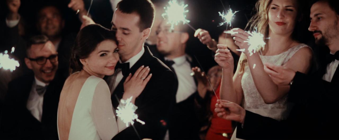 Para młoda otoczona gośćmi weselnymi z zimnymi ogniami, uśmiechnięta panna młoda w sukni z odkrytymi plecami i bukiecikiem na nadgarstku, pan młody w czarnym smokingu z muszką.