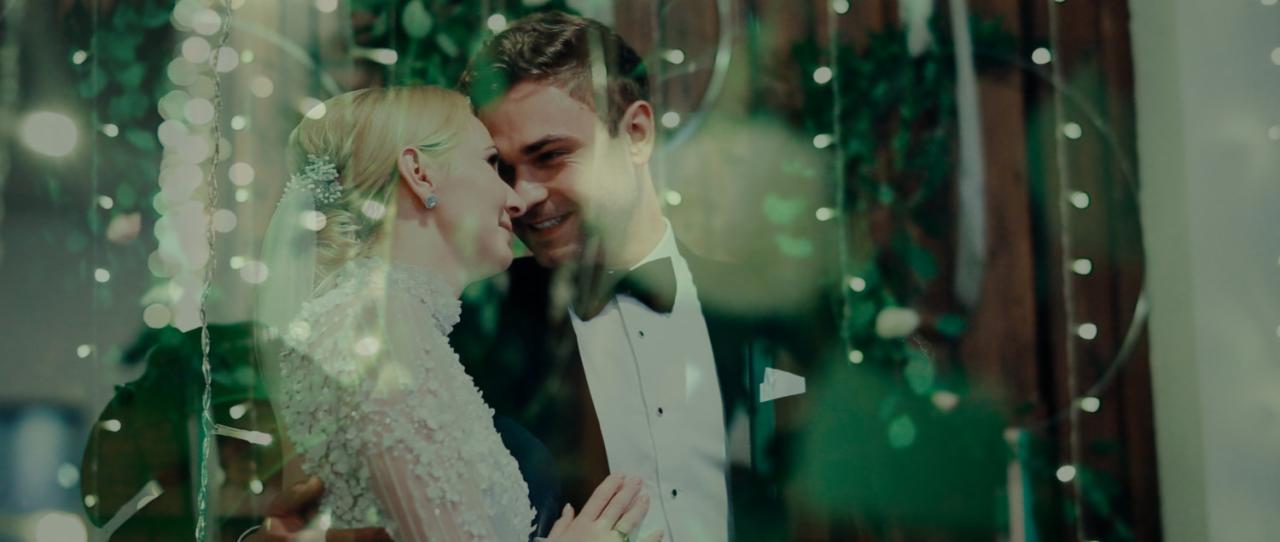 Uśmiechnięta para młoda, panna młoda w koronkowej sukni z kwiatowymi aplikacjami i delikatnym upięciu, pan młody w smokingu, w tle dekoracja z zielonych gałązek i światełek, ujęcie przez szybę.