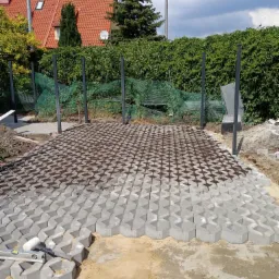 Układanie ażurowej kostki brukowej na piaskowym podłożu, widoczne narzędzia: poziomica i kielnia, w tle metalowe słupki przyszłego ogrodzenia i dom z czerwoną dachówką.