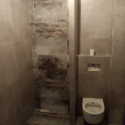 Wykończona łazienka z podwieszaną toaletą, szare płytki imitujące kamień na ścianach i podłodze, widoczne przyłącza do baterii.