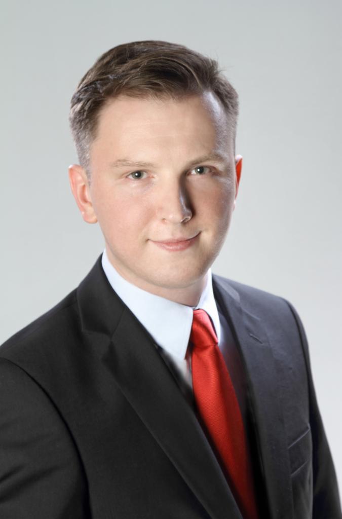 Portret mężczyzny w czarnym garniturze i czerwonym krawacie na jasnoszarym tle.