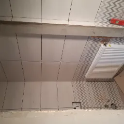 Łazienka w trakcie układania płytek: białe płytki kwadratowe na ścianie, częściowo ułożone płytki z wzorem w zygzaki, widoczny grzejnik i poziomica.