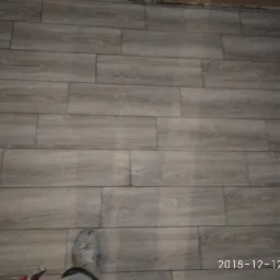 Ułożone szare panele podłogowe imitujące drewno, widoczny fragment buta i data na zdjęciu.