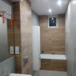 Nowoczesna łazienka z drewnopodobnymi płytkami na ścianach, białą zabudową wnęki i wanną. Widoczne detale: chromowana bateria, szklana kabina prysznicowa z czerwonymi akcentami, minimalistyczne...