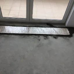 Wpuszczony w posadzkę grzejnik kanałowy, zabezpieczony folią ochronną, w trakcie montażu przy oknie z widokiem na taras.