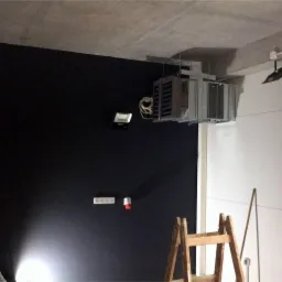 Szare urządzenie klimatyzacyjne zamontowane pod sufitem w pomieszczeniu z czarną ścianą, widoczne również oświetlenie i drabina.