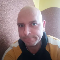 Portret mężczyzny z ogoloną głową, ubranego w bluzę z kapturem w kolorach czarnym, białym i żółtym, na tle korkowej ściany z elementami dekoracyjnymi.