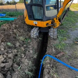 Żółta mini koparka JCB wykonuje wąski wykop w ziemi, w którym ułożona jest niebieska rura karbowana. W tle widoczne zielone trawy i fragment ogrodzenia.