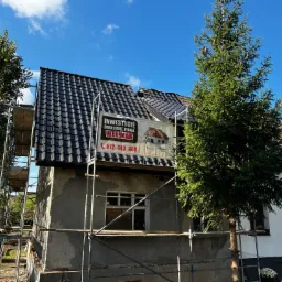 Dom w trakcie remontu dachu z częściowo położoną nową dachówką, widoczne rusztowania i baner reklamowy firmy budowlanej.