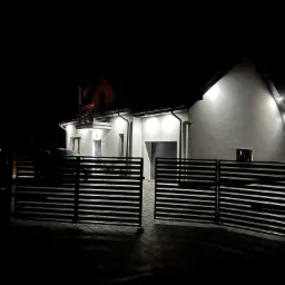 Nowoczesny, oświetlony dom jednorodzinny z płaskim dachem, widoczny nocą przez otwartą, poziomą bramę wjazdową. Na podjeździe stoi samochód. Oświetlenie elewacyjne podkreśla białą fasadę.