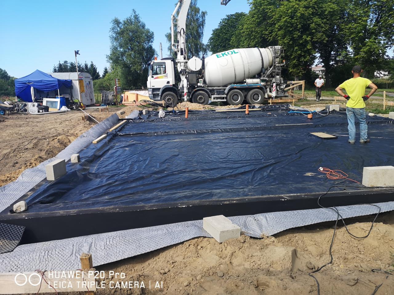 Przygotowanie pod fundamenty: czarna folia na podłożu, betonowa rama, w tle betoniarka EKOL i pracownicy na placu budowy.