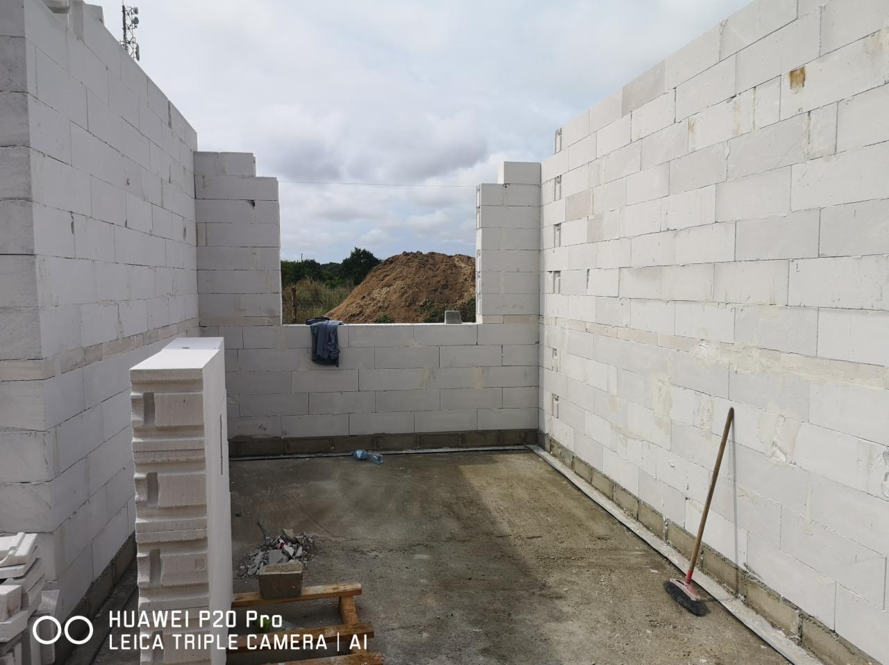 Wnętrze surowego domu w trakcie budowy, ściany wzniesione z białych bloczków, widoczne okno z widokiem na hałdę ziemi, na podłodze resztki materiałów i miotła oparta o ścianę.