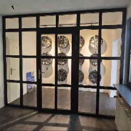 Przeszklone, czarne drzwi z metalowymi szprosami, widok na wnętrze z ekspozycją felg samochodowych. Po prawej stronie okno z czarną ramą.