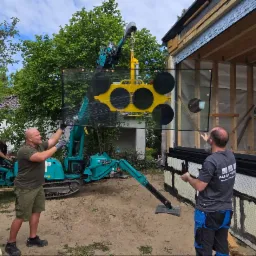 Montaż okna: Ekipa budowlana instaluje duże okno za pomocą mini żurawia przyssawkowego na tle zieleni. Widoczne elementy konstrukcji budynku.