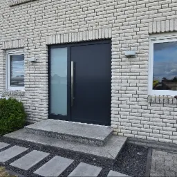 Nowoczesne, antracytowe drzwi wejściowe z długim uchwytem i oknem bocznym, w Chojnie. Biała cegła elewacyjna i okno PCV w tle. Ścieżka z płyt.
