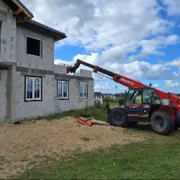 Montaż okien PCV w surowym budynku z pomocą podnośnika teleskopowego Maniou. Widoczne białe okna i otwory okienne bez szyb. Niebieskie niebo z chmurami w tle.