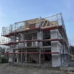 Wznoszenie dwukondygnacyjnego domu z białych bloczków, widoczne rusztowania i drewniana konstrukcja dachu. Brak okien i drzwi. Etap budowy w surowym stanie.