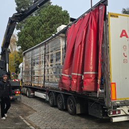 WINDOLUX Przemysław Wojciechowski - Rozładunek okien PCV z ciężarówki za pomocą dźwigu. Pracownicy w odblaskowych kamizelkach zabezpieczają ładunek. Widoczny fragment budynku w tle.