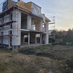 WINDOLUX Przemysław Wojciechowski - Budowa domu w stanie surowym otwartym, z rusztowaniami i widoczną konstrukcją dachu. Osoba stoi na działce. Słońce w tle.