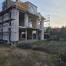 Budowa domu w stanie surowym otwartym, z rusztowaniami i widoczną konstrukcją dachu. Osoba stoi na działce. Słońce w tle.