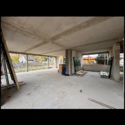 Surowy stan wnętrza budynku w trakcie budowy. Widoczne betonowe ściany, słupy, strop i przygotowane do montażu okna. Na zewnątrz widoczny krajobraz.