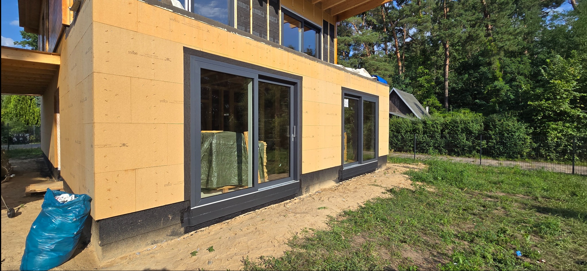 Dom w trakcie budowy, widoczne okna przesuwne z szarymi ramami, ściany z płyt izolacyjnych. Na pierwszym planie niebieski worek na śmieci.