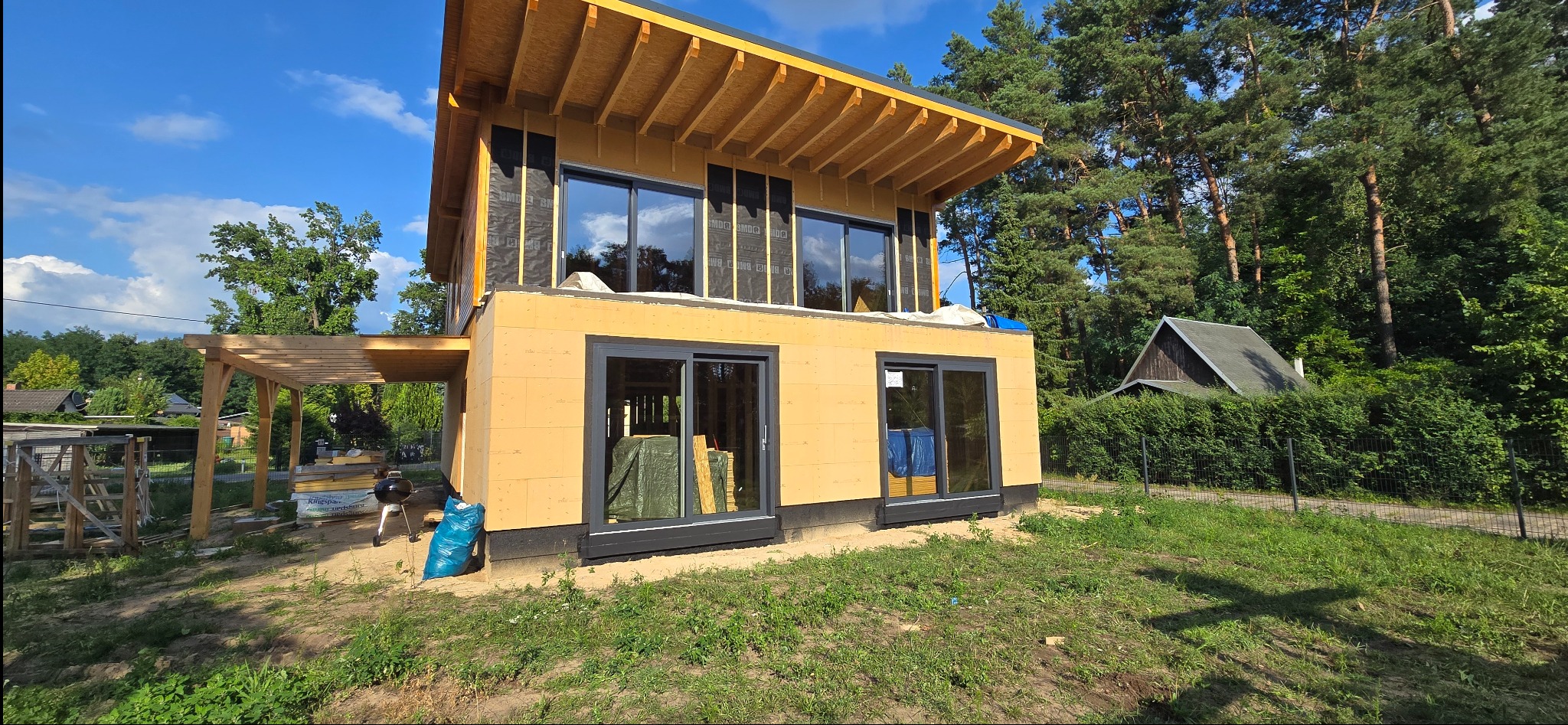 Surowy stan domu z zamontowanymi oknami z czarnymi ramami, widoczne drewniane elementy konstrukcyjne i izolacja. Dom w otoczeniu drzew.