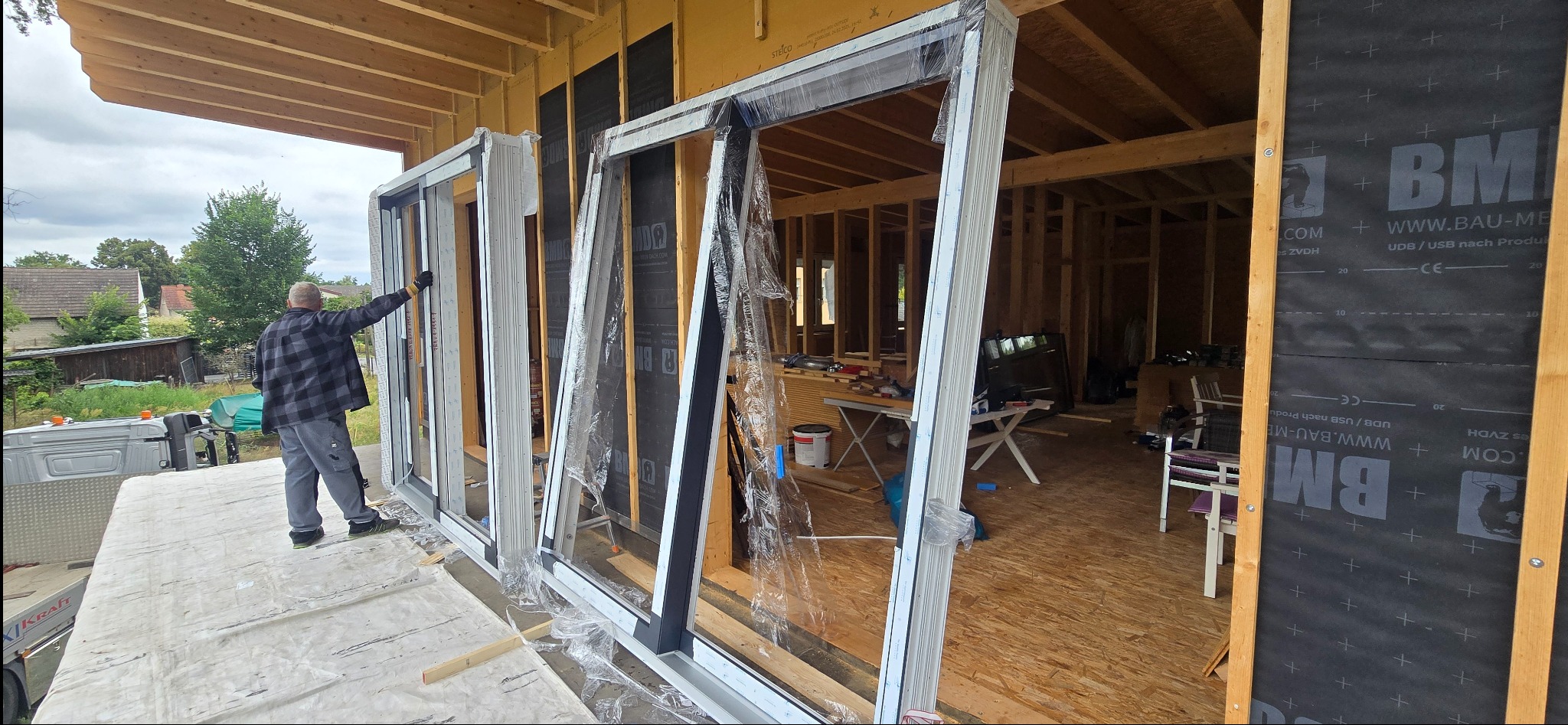 Montaż okien w surowym stanie budynku. Widoczne okna PCV z białymi ramami, częściowo jeszcze w folii ochronnej. Pracownik w tle. Budynek w trakcie budowy.