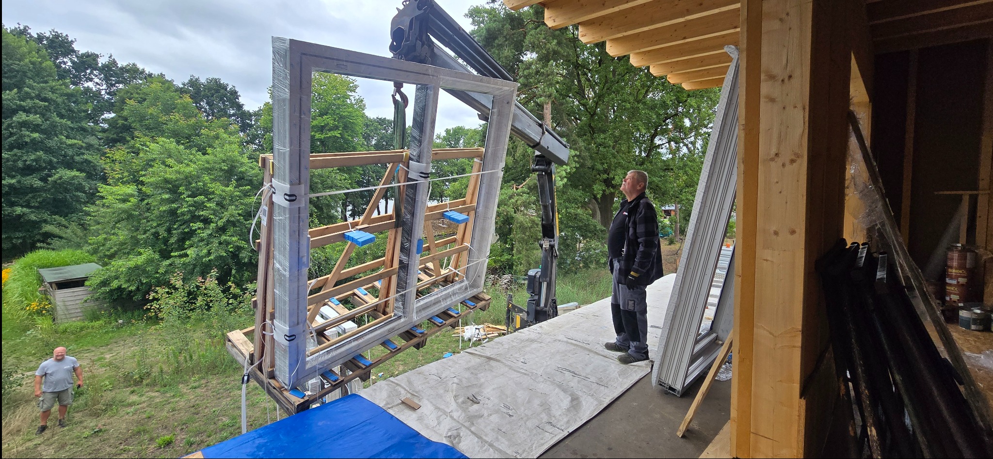 Montaż okien z użyciem dźwigu w Chojnie. Widoczne okna PCV w folii ochronnej, pracownik nadzorujący i fragment konstrukcji budynku w trakcie budowy.