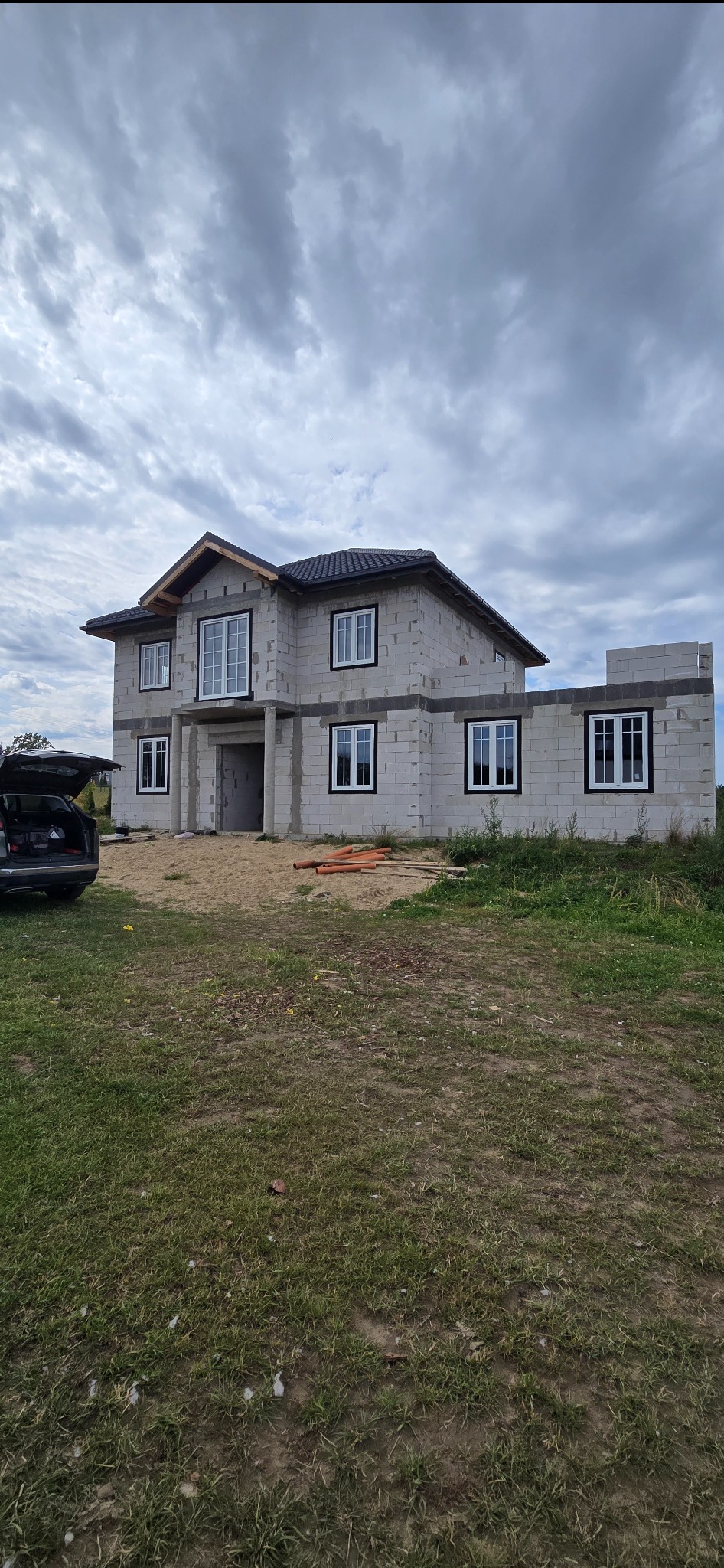 Surowy stan domu w budowie z czarnymi ramami okiennymi i białymi oknami, widok z zewnątrz, częściowo otwarty bagażnik samochodu po lewej stronie.