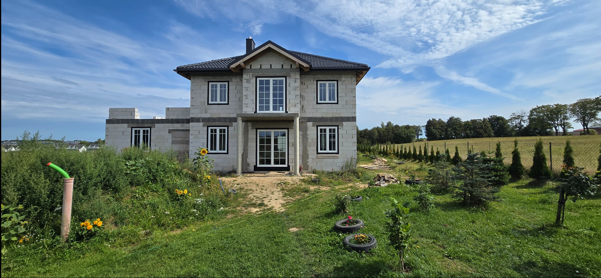 Dom w trakcie budowy z zamontowanymi oknami i drzwiami z czarnymi ramami. Widoczny surowy stan ścian i zagospodarowany teren wokół budynku.