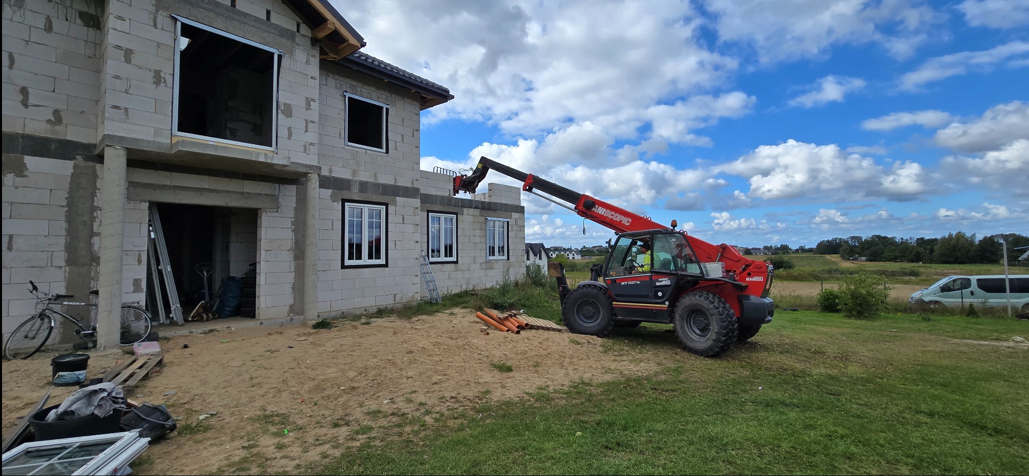 Montaż okien PCV w surowym budynku z pomocą podnośnika teleskopowego Maniou. Widoczne białe okna i otwory okienne bez szyb. Niebieskie niebo z chmurami w tle.