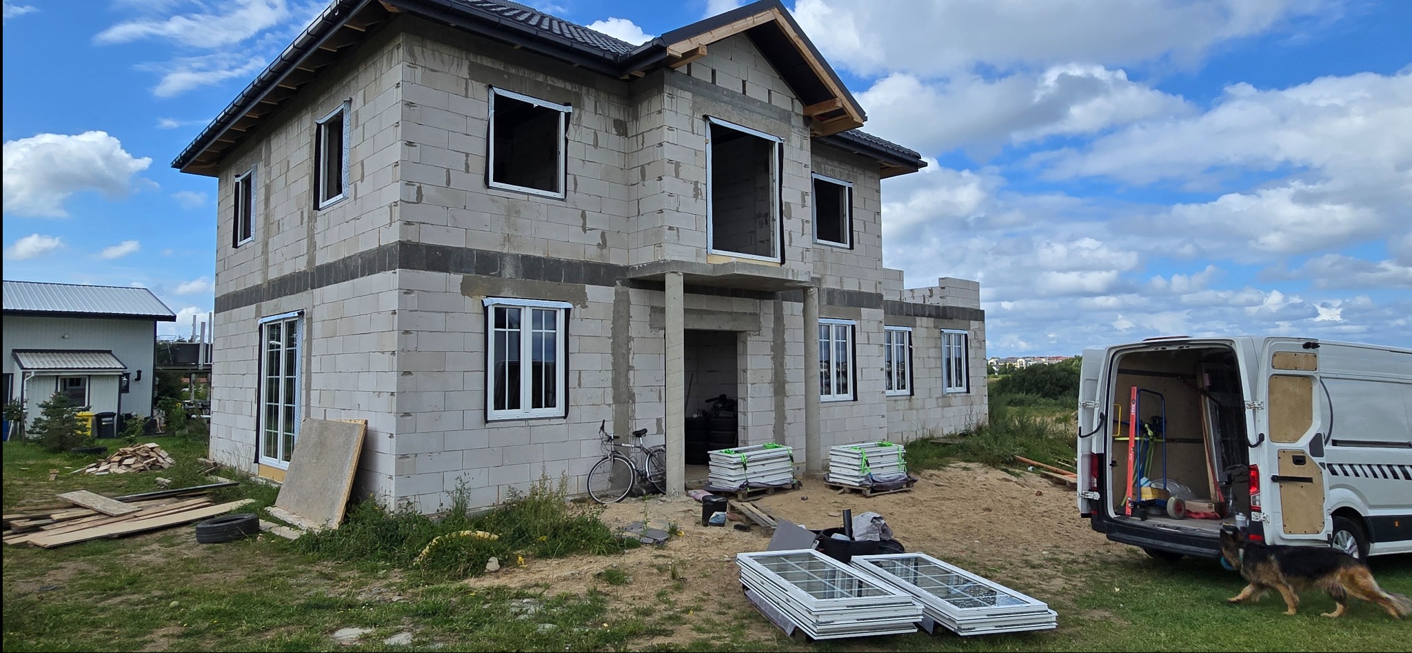 Dom w trakcie budowy z zamontowanymi oknami PCV. Na pierwszym planie okna czekające na montaż i samochód dostawczy z narzędziami.