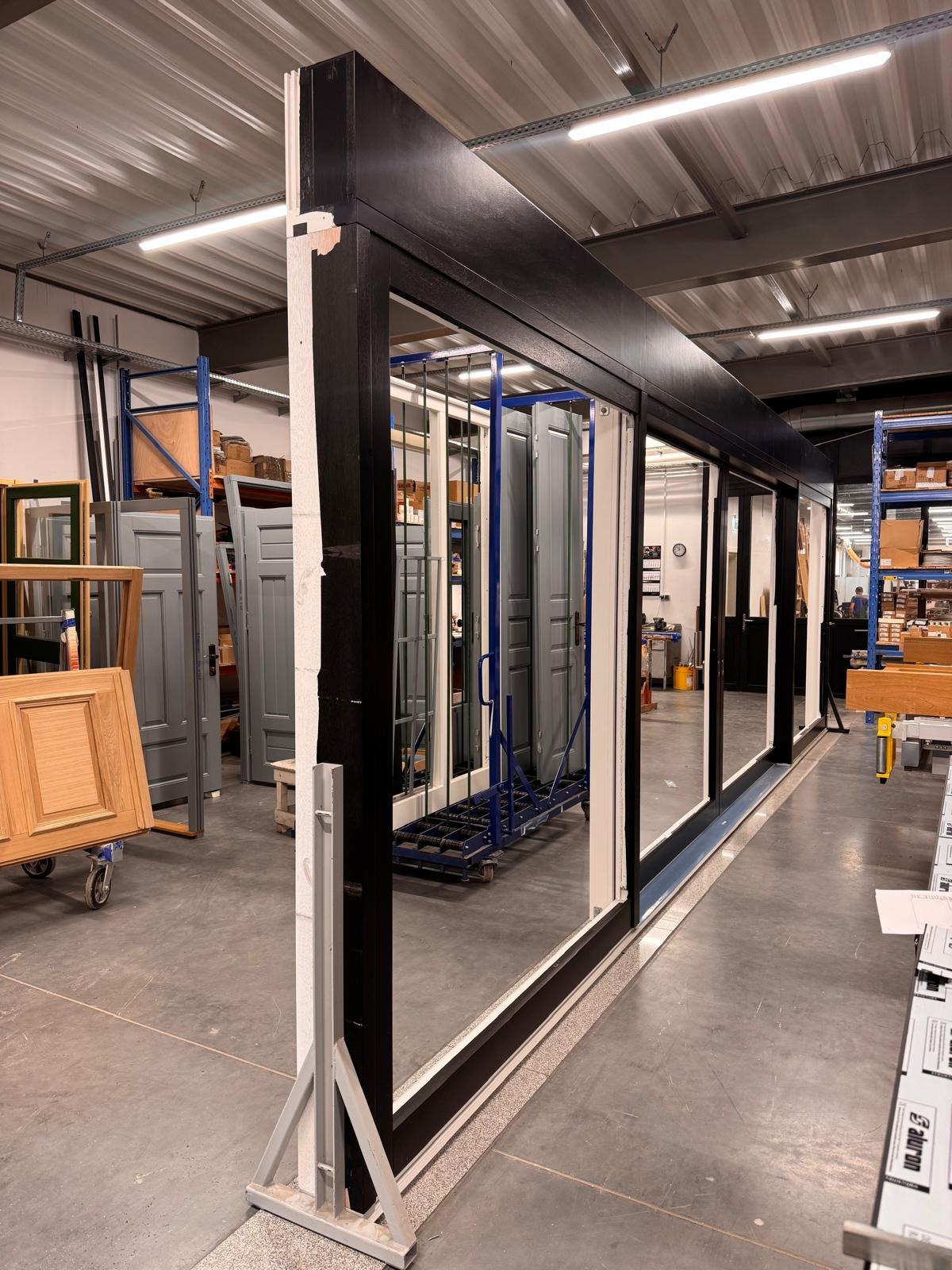 Wystawa stolarki okiennej w hali produkcyjnej w Chojnie. Widoczne ramy okienne i drzwi na wózkach transportowych. Oświetlenie przemysłowe.