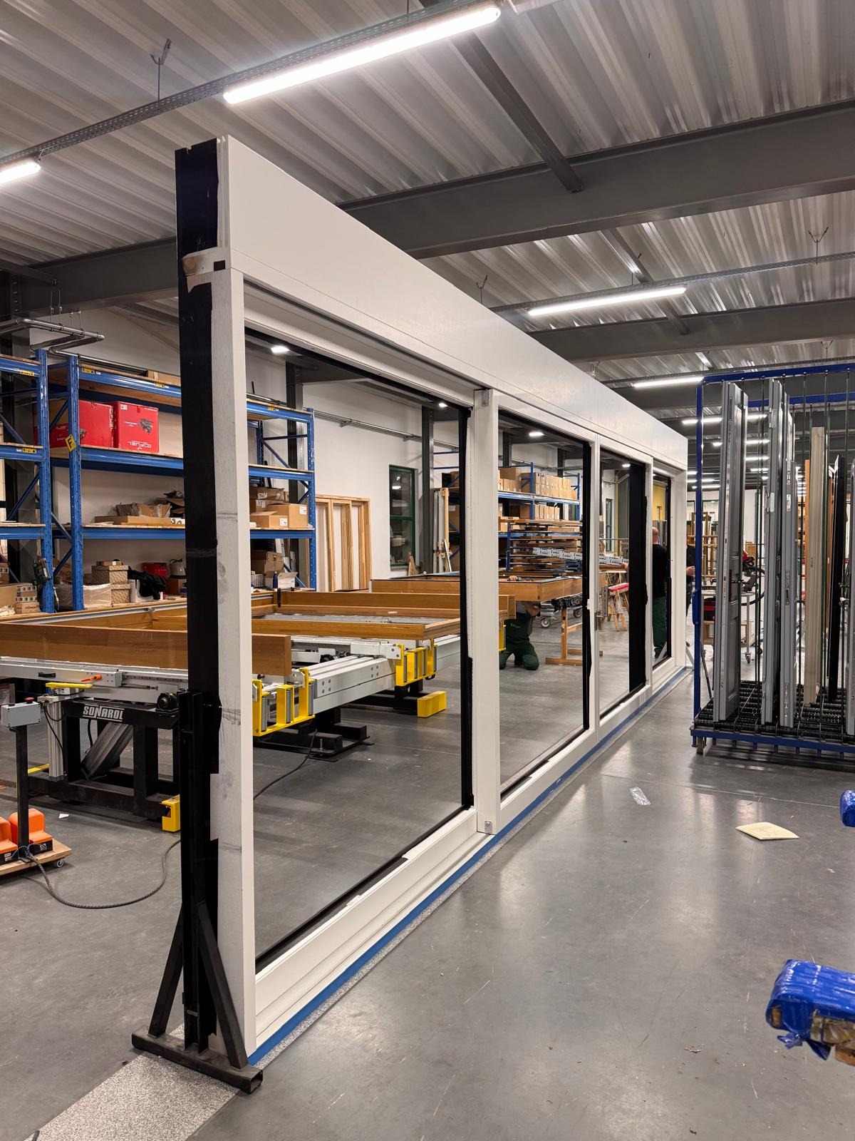 Długie, białe okno przesuwne z czarnymi ramami, ustawione w hali produkcyjnej z regałami i maszynami w tle. Widoczne detale konstrukcyjne i elementy montażowe.