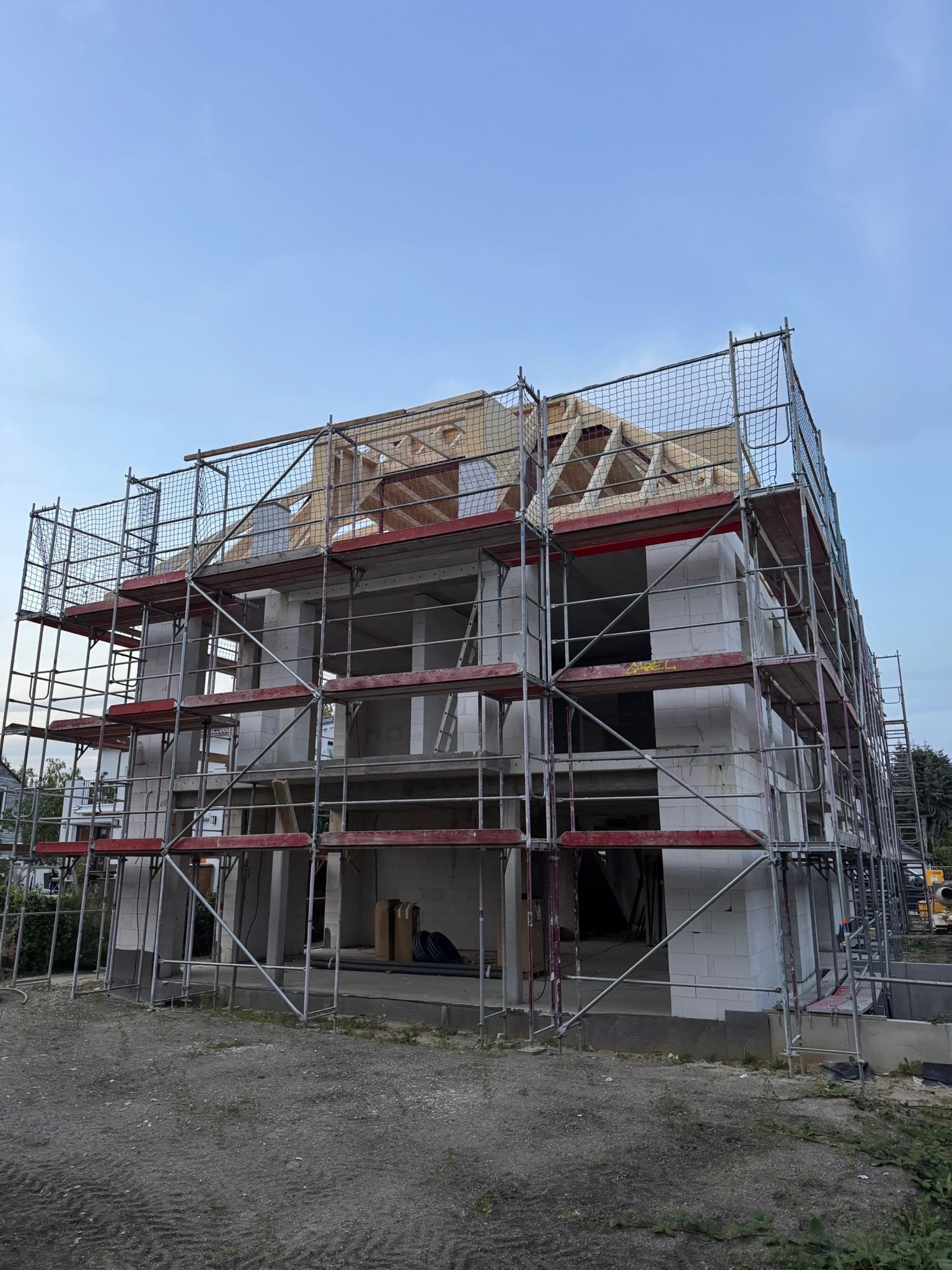 Wznoszenie dwukondygnacyjnego domu z białych bloczków, widoczne rusztowania i drewniana konstrukcja dachu. Brak okien i drzwi. Etap budowy w surowym stanie.