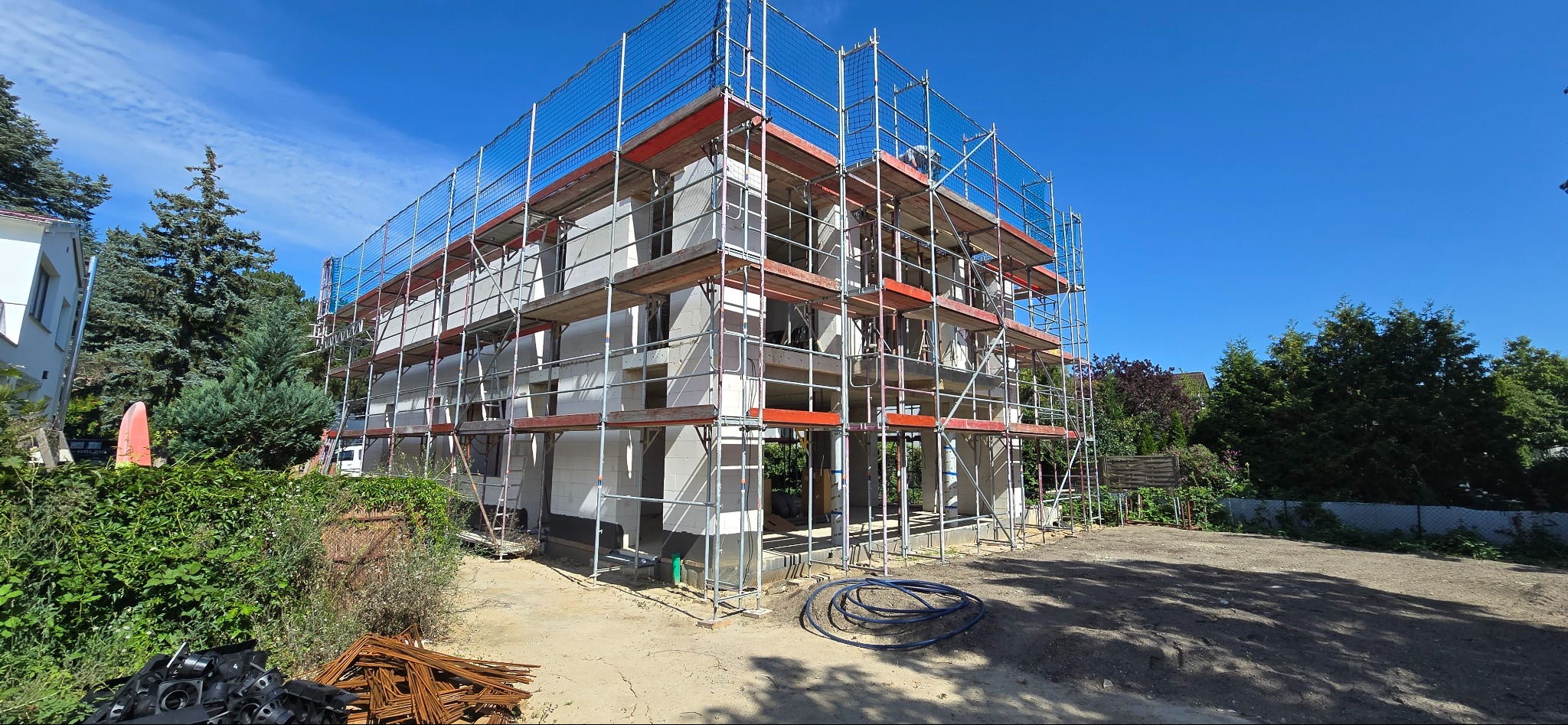 Budowa domu z białych bloczków, otoczona rusztowaniami i siatką ochronną. Widoczne materiały budowlane i niebieskie węże. Słoneczny dzień, błękitne niebo.