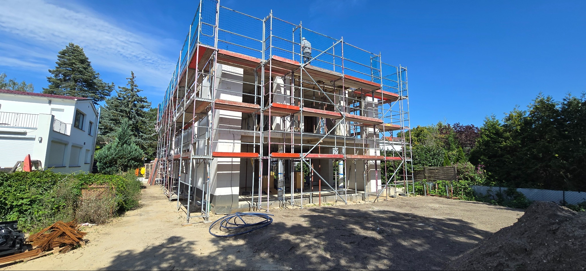 Budynek w trakcie budowy, otoczony rusztowaniem. Widoczne białe bloczki, niebieskie niebo i drzewa w tle. Na rusztowaniu stoi osoba w jasnym ubraniu.