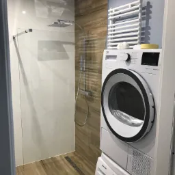 Nowoczesna łazienka z kabiną prysznicową typu walk-in, wykończona płytkami imitującymi drewno, z pralką i suszarką w słupku oraz grzejnikiem drabinkowym.