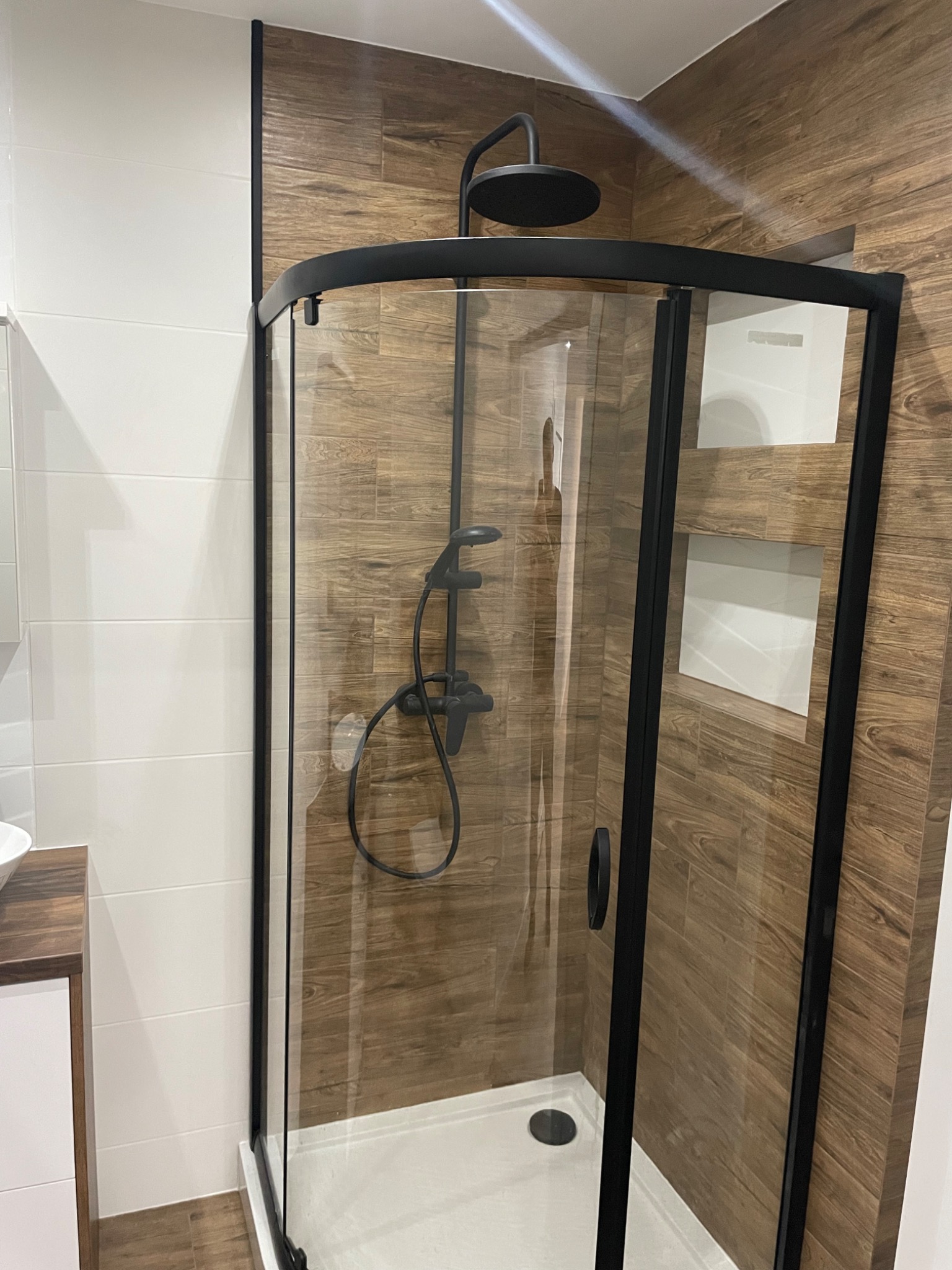 Nowoczesna kabina prysznicowa z czarną armaturą i ramą, na ścianach płytki imitujące drewno oraz białe płytki.