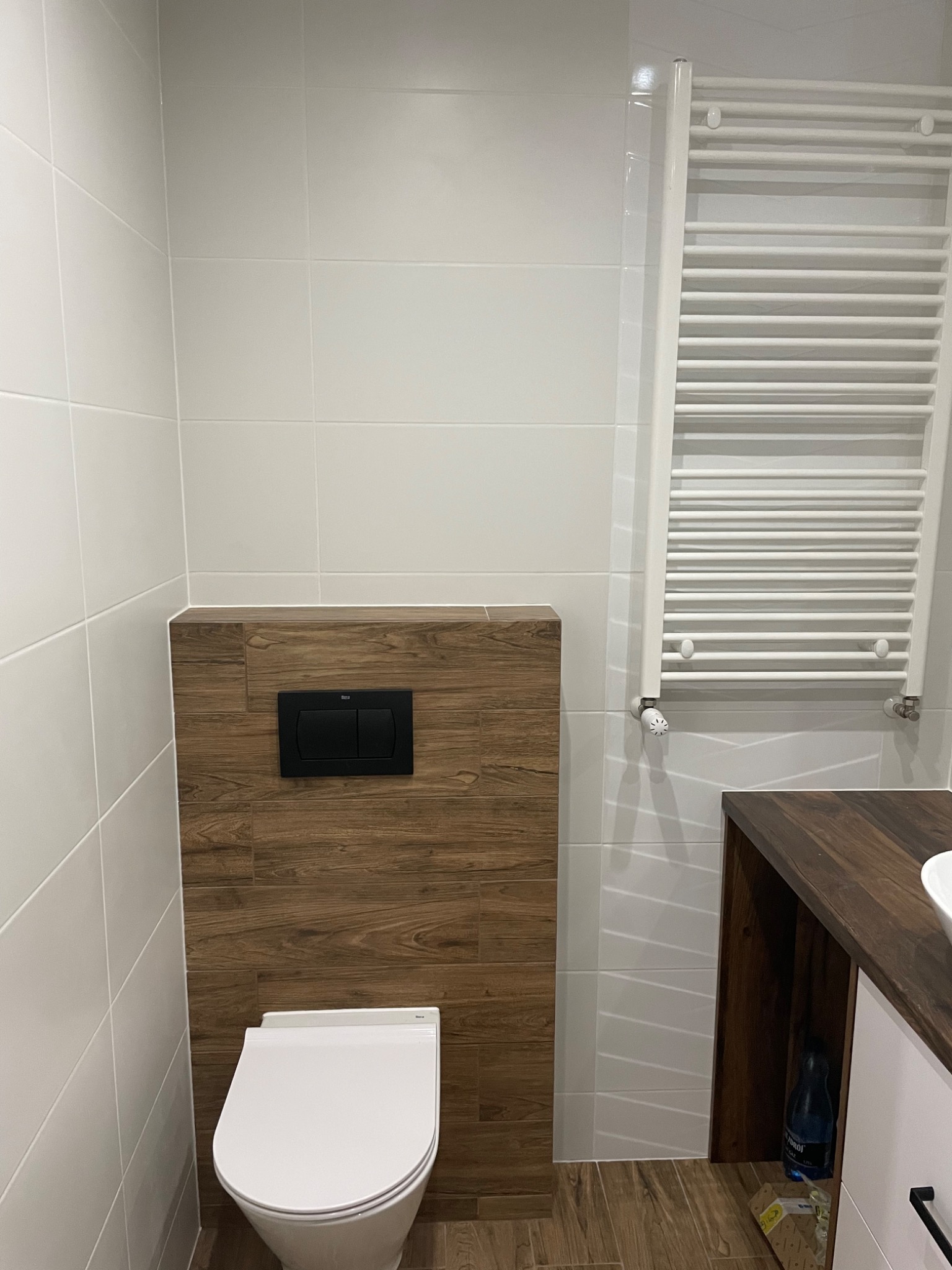 Nowoczesna łazienka z białymi płytkami, zabudowaną toaletą z drewnopodobną obudową i grzejnikiem drabinkowym.