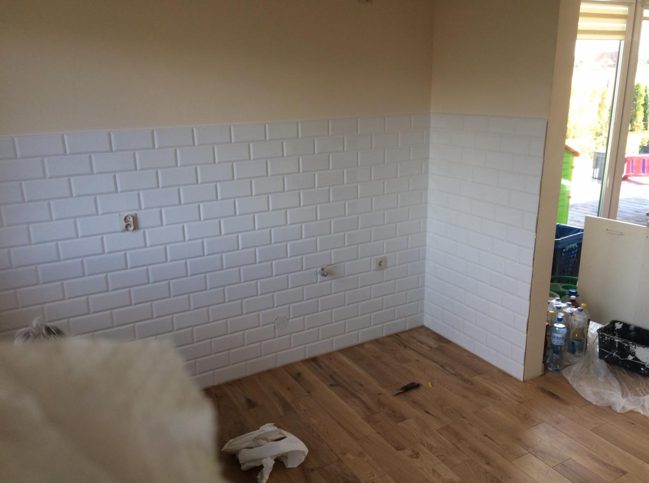 Ściana w kuchni wyłożona białymi, błyszczącymi kafelkami typu metro, widoczne gniazdka elektryczne i fragment drewnianej podłogi z resztkami materiałów budowlanych i narzędziami, okno z widokiem...