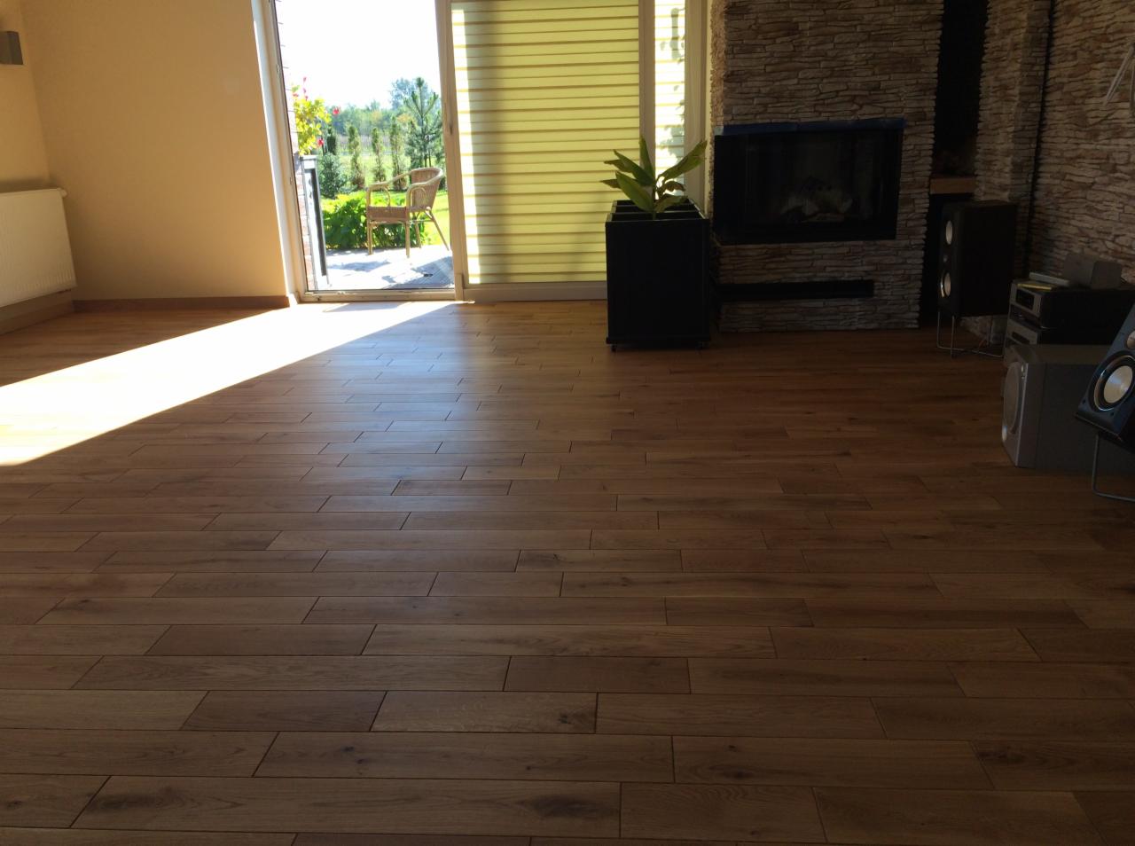 Ułożone panele podłogowe w salonie z widokiem na ogród, jasne światło słoneczne pada na podłogę, w tle kominek z kamienia i sprzęt audio.