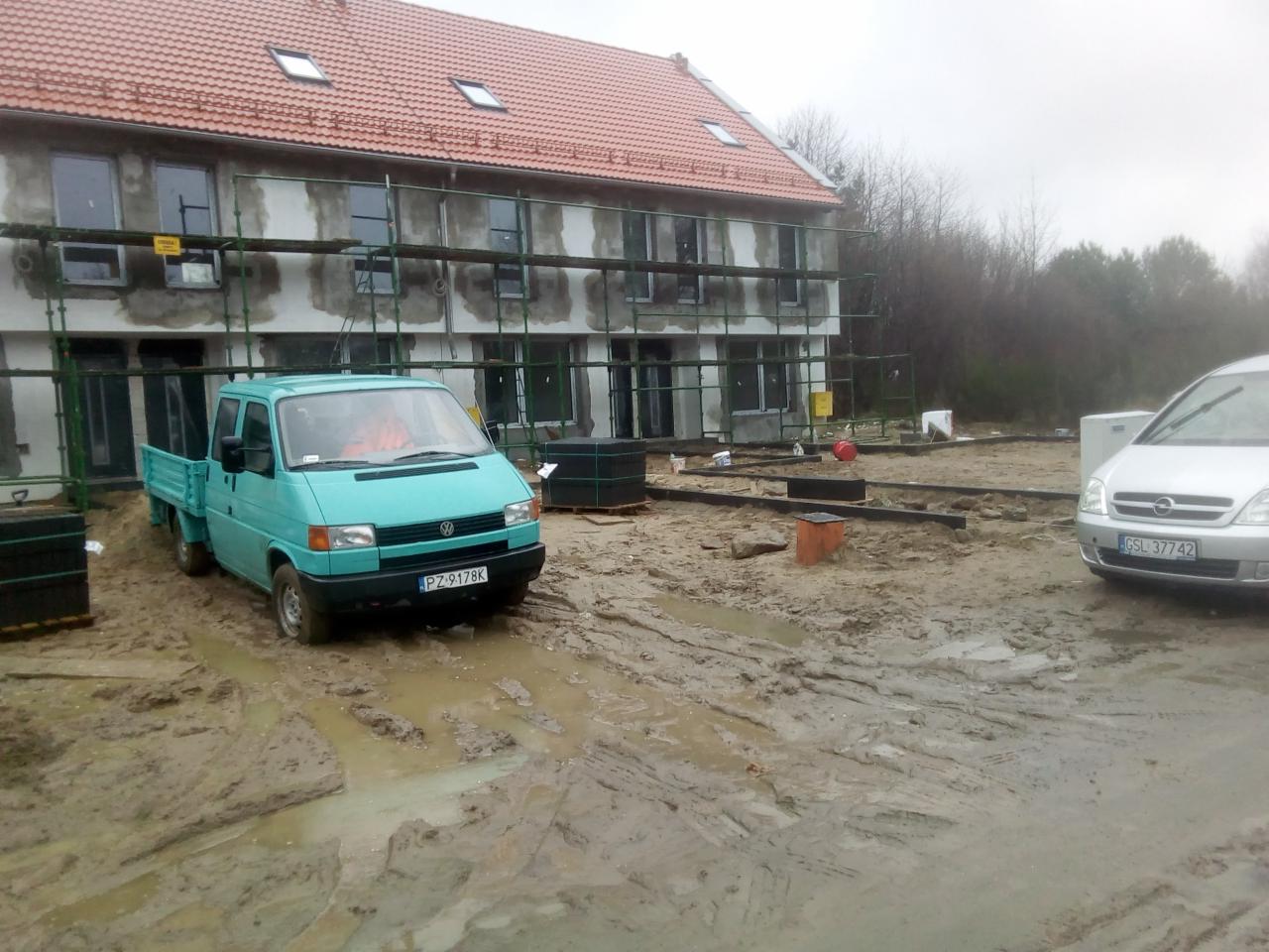 Turkusowy pick-up Volkswagen z kostką brukową na pace stoi na błotnistym placu budowy przed budynkiem w trakcie remontu, w tle widoczne rusztowania.