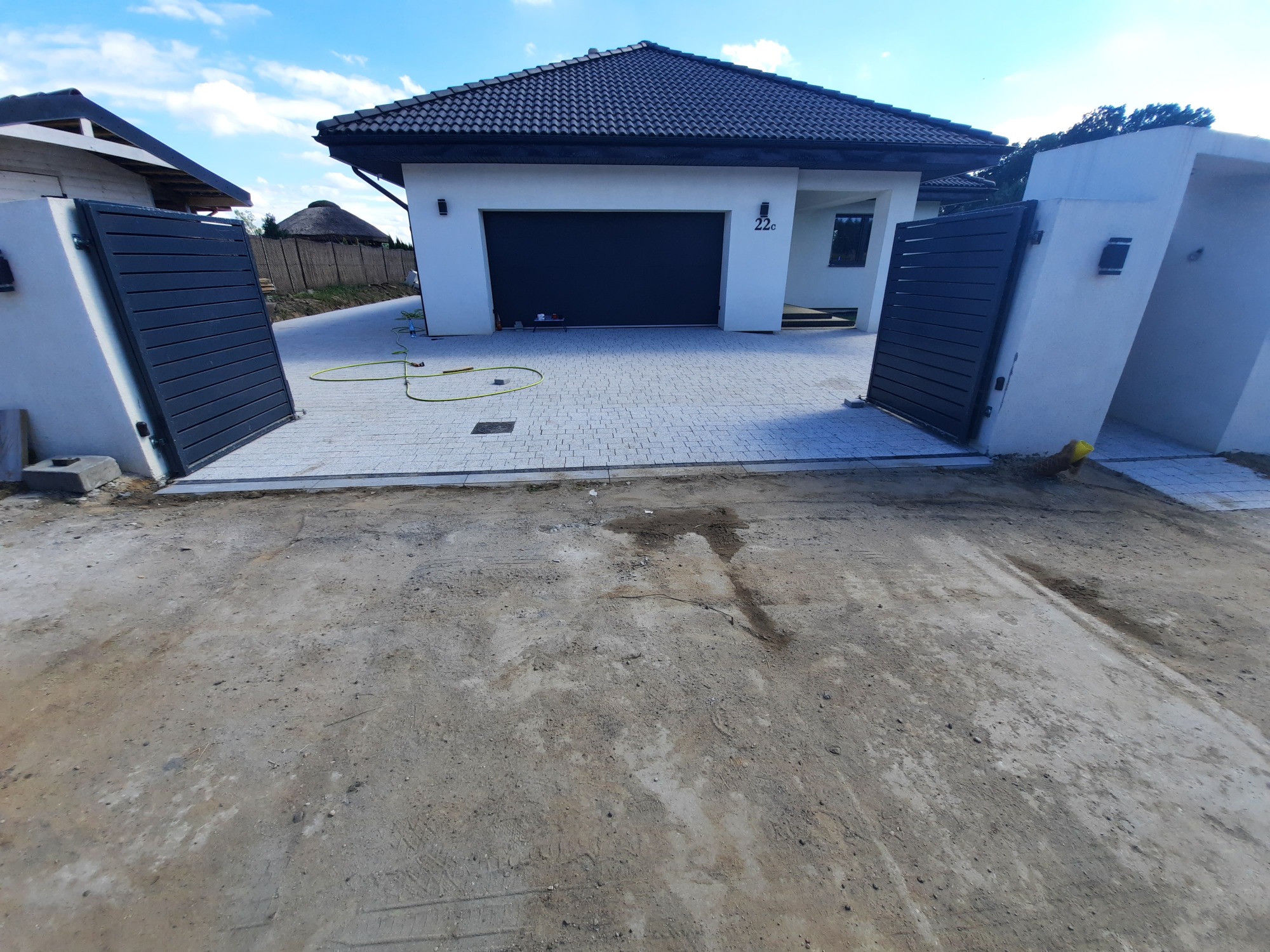 Nowoczesny podjazd z jasnoszarej kostki brukowej prowadzący do domu z garażem i automatyczną bramą wjazdową; widoczny fragment trawnika i niezakończone prace ziemne.